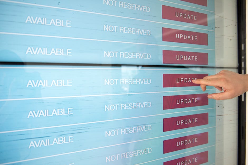 A close-up view of a digital booking display board with multiple rows showing 'AVAILABLE' and 'NOT RESERVED' statuses, alongside red 'UPDATE' buttons. A person's hand with a light skin tone is pressin