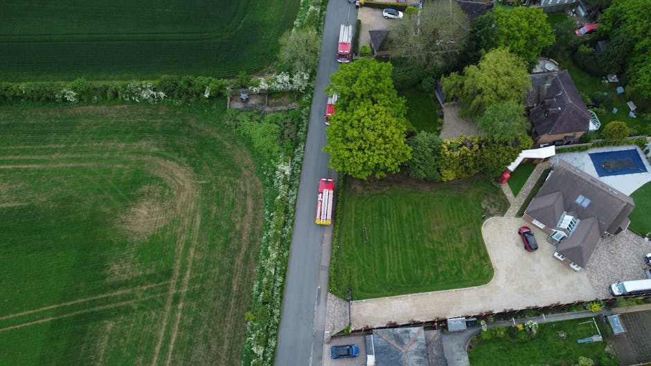 An aerial view of a suburban area showing a well-maintained garden along a residential street. On the right, there is a modern house with a grey roof, a driveway with a red car parked, and a backyard